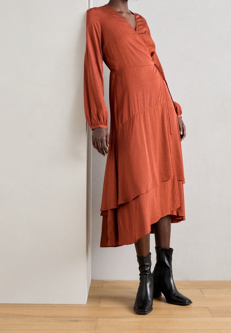 Femme portant une robe midi à manches longues couleur rouille, avec un ourlet en couches, et des bottes à talons jusqu'au mollet noires, se tenant contre un mur simple.