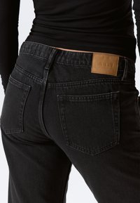 Jeans in denim nero con vita alta, due tasche posteriori e un'etichetta in pelle marrone con il marchio "WEEKDAY" sulla vita.