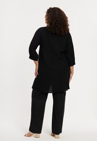 Zwarte geribbelde tuniek met driekwart mouwen, iets langere achterkant, gecombineerd met losse zwarte broeken. Eenvoudig ontwerp, gestructureerde stof.