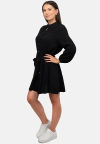 Abito nero con bottoni, vita cinturata, maniche lunghe e tessuto texturizzato. Indossato con sneakers bianche.