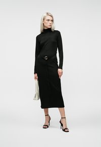 Čierne dlhé šaty s dlhým rukávom a vysokým výstrihom, s opaskom so zlatým akcentom. Kombinované s čiernymi sandálmi na vysokom podpätku a bielou kabelkou.