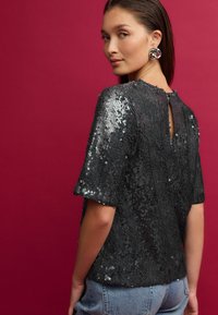 Haut à sequins noirs avec des manches courtes, surface texturée et détail en forme de trou de serrure à l'arrière, associé à un jean bleu clair sur un fond rose.