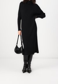 Robe noire longue côtelée à col montant, portée avec des bottes noires ajustées à bouts pointus et détails argentés. Comprend un petit sac à main noir.