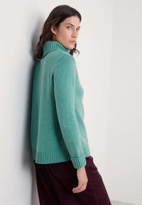 Tirkizna pletena dukserica s rebrastim visokim ovratnikom, dugim rukavima i rebrastim rubom, u kombinaciji s bordo odrezanim hlačama. Glatka tekstura.