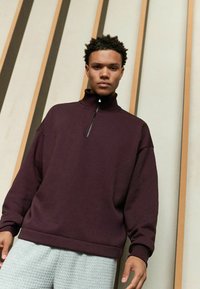 Burgunderfarbener Sweatshirt mit Reißverschluss am Hals, mit gerippten Bündchen und Saum, im lässigen Schnitt. Getragen mit hellgrauen strukturierten Hosen.