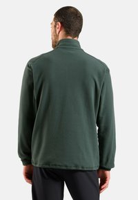 Homme portant une veste en polaire vert foncé et un pantalon noir, se tenant de dos face à la caméra devant un fond uni.