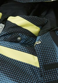 Mehrfarbige Jacke mit einem blauen und schwarzen gepunkteten Muster und gelben Akzenten. Sie hat Druckknöpfe und einen strukturierten Stoff.