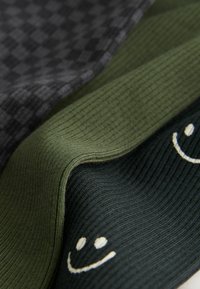 Trei straturi de țesătură: model în carouri negre, textură ribbed verde măsliniu, și verde închis cu detalii cu fața zâmbitoare albă.