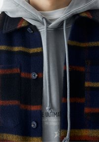 Giacca di flanella a quadri in navy, arancione e giallo con chiusura a bottoni; indossata sopra un hoodie grigio con stampa grafica e cordino.