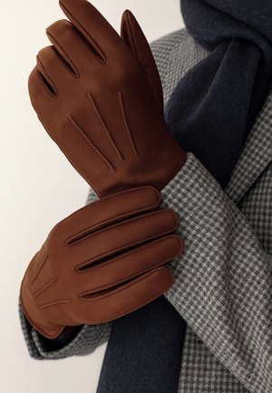 Brown leather gloves with stitch detailing, held together, against a grey plaid coat and a dark blue scarf background.