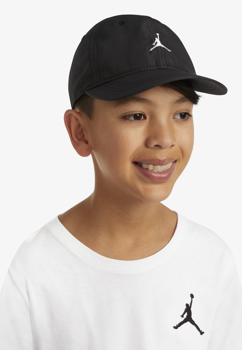 Černá baseballová čepice s bílým logem Jumpman. Vyrobena z měkkého materiálu, má zakřivený kšilt a nastavitelný pásek. Nošena s bílým tričkem.