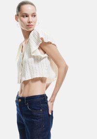 Weißes, kurz geschnittenes Strickoberteil mit strukturiertem Muster und gerafften Ärmeln, kombiniert mit dunkelblauen, hochtaillierten Jeans mit Standardbeschlägen.