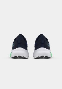 Marineblå joggesko med mesh-overdel, hvit såle og grønne detaljer på hælen. Har en teksturert såle for grep og et stoffhekle.