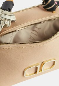 Borsa in tessuto beige con accessori in oro, chiusura con zip e una sciarpa decorativa annodata al manico. Interno con rivestimento liscio.