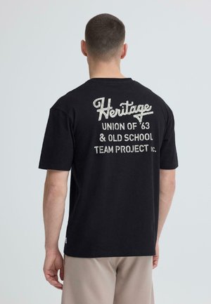 Homme portant un T-shirt noir avec un texte blanc « Heritage Union of '63 & Old School Team Project » dans le dos, pantalon beige, fond clair uni.