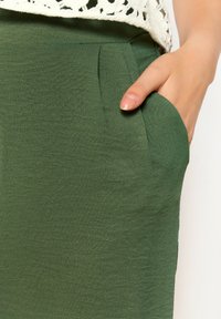 Green fabric with a smooth texture featuring a side pocket. The design includes pleats at the waistband and a knitted white top overlay.