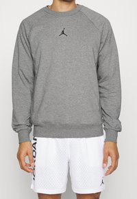 Felpa grigia con maniche lunghe e polsini a coste, caratterizzata da un logo Jumpman nero sul petto. Indossata con pantaloni corti in rete bianchi.