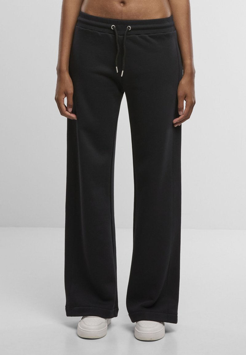 Black sweatpants with wide legs, elastic waistband, and drawstring detail. Soft texture, minimal design, paired with white sneakers.