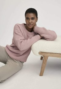 Rosa gestrickter Pullover mit rundem Halsausschnitt und gerippten Bündchen, kombiniert mit hellgrauen Hosen. Weiße, strukturierte gepolsterte Couch mit Holzbeinen.