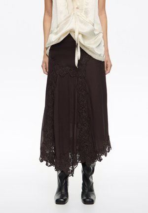 Mujer vistiendo una falda midi marrón oscuro con borde de encaje y botas de cuero negras, combinada con una blusa color crema con un nudo en el frente.