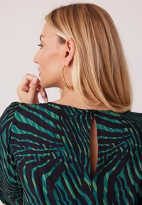 Czarny top w zielono-błękitnym paskowanym wzorze, okrągły dekolt, małe rozcięcie z tyłu i miękka tkanina. Widoczne są duże złote kolczyki w kształcie koła.