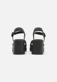 Svart, høyhælte sandaler med tykk klossjede, teksturerte skinnremmer og justerbar ankelspenne for sikker passform.