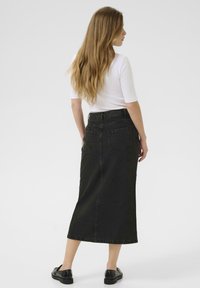 Jupe midi en denim noir avec taille haute, deux poches arrière et une texture lisse, assortie à un top blanc ajusté et des chaussures noires.