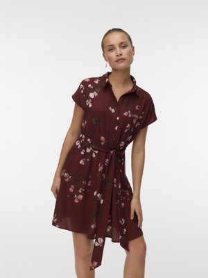 Femme portant une robe fleurie bordeaux à manches courtes, avec un col et une ceinture à la taille, debout devant un fond blanc uni.