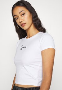 T-shirt blanc court en coton, avec des manches courtes et un logo script noir à l'avant. Texture lisse, style ajusté.