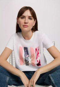 Biała bawełniana koszulka z abstrakcyjnym wzorem kwiatu w odcieniach różu, granatu i złota. Model w zrelaksowanej pozie, w zestawieniu z niebieskimi dżinsami.