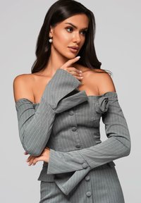 Szara bluzka typu off-shoulder w pionowe paski, z dużym detalem w postaci kokardy na ramieniu i rozkloszowanymi rękawami. Posiada zapięcie na guziki i strukturalną tkaninę.
