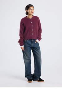 Burgundy knitted cardigan with six button front, ribbed texture, and balloon sleeves paired with loose-fitting dark blue jeans and black shoes.