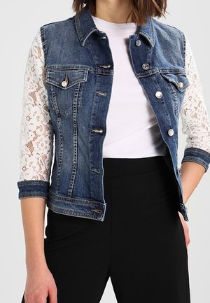 Denim vest with lace white sleeves worn over a white top and paired with black high-waist pants.