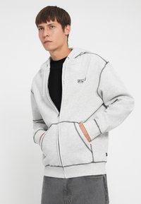 Grå zip-up hettegenser laget av mykt stoff, med svarte sømdetaljer og lommer. Brukt over en svart skjorte, med hette og avslappet passform.