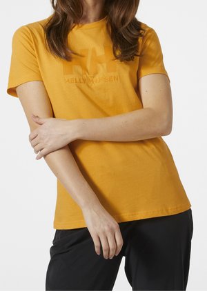 Žena oblečená v hořčicově žlutém tričku s krátkým rukávem značky Helly Hansen a černých kalhotách, s překříženýma rukama v pase.