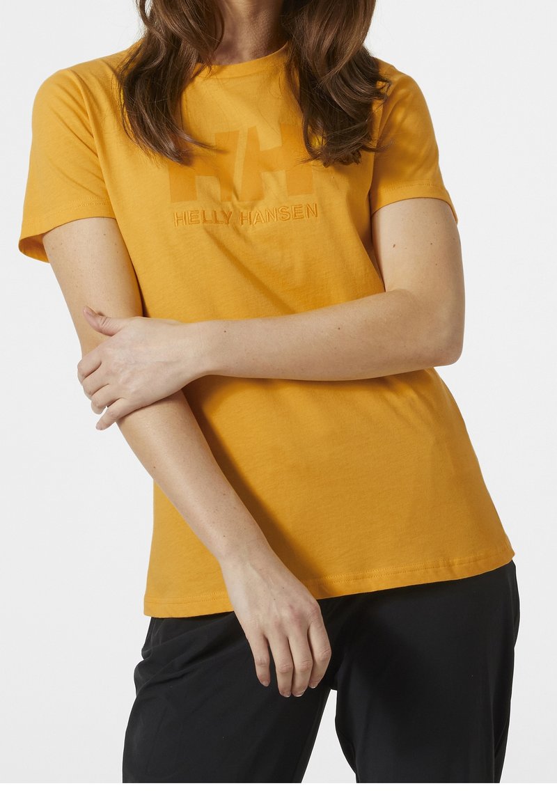 Femme portant un T-shirt Helly Hansen à manches courtes jaune moutarde et un pantalon noir, les bras croisés à la taille.