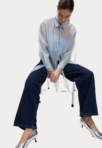 Blusa trasparente azzurro chiaro con colletto e bottoni gioiello sul davanti, abbinata a jeans a gamba larga blu scuro e scarpe con tacco argento.