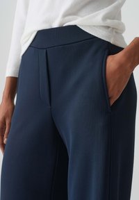 Marineblauwe, wijde broek met een platte tailleband, voorzien van zijzakken en voorplooien. Het materiaal lijkt glad en gestructureerd.