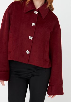 Femme portant une veste courte en laine bordeaux avec de gros boutons argentés et des manches larges, associée à un pantalon noir, sur un fond uni.