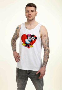 Camiseta sin mangas blanca con un gráfico de Mickey y Minnie Mouse sobre un fondo de corazón rojo. La modelo tiene tatuajes y lleva jeans grises.