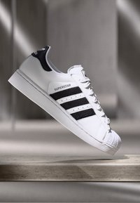 Biele tenisky Adidas Superstar s čiernymi pruhmi a pätou, vznášajúce sa nad kamenným povrchom v jemnom, neutrálnom osvetlení.