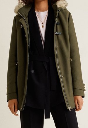 Une personne porte une parka vert olive avec une capuche en fourrure, un blazer noir ceinturé, une chemise blanche, un pantalon noir et des colliers en or superposés.