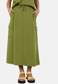 Green maxi skirt with an elastic waistband and drawstring. Features two side pockets; paired with yellow sandals on a white background.