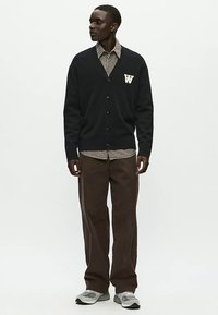 Marineblauer Cardigan mit weißem "W"-Logo und Knopfverschluss, getragen über einem gestreiften Hemd. Kombiniert mit brauner Hose und grauen Turnschuhen.