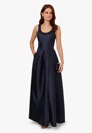 Granatowa satynowa sukienka wieczorowa z wyszywaną okrągłą szyją, sylwetką w kształcie litery A oraz kieszeniami bocznymi. Modelka trzyma małą kopertówkę i ma na sobie buty z odkrytymi palcami.
