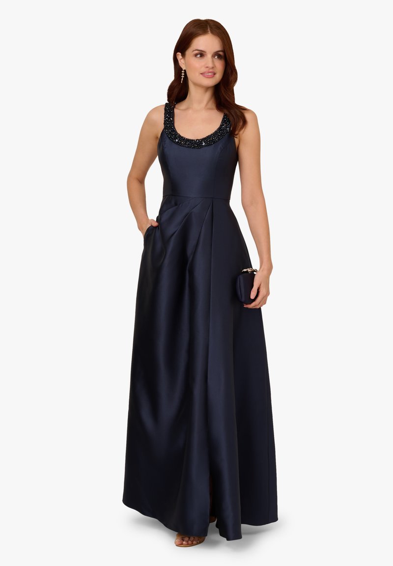 Granatowa satynowa sukienka wieczorowa z wyszywaną okrągłą szyją, sylwetką w kształcie litery A oraz kieszeniami bocznymi. Modelka trzyma małą kopertówkę i ma na sobie buty z odkrytymi palcami.