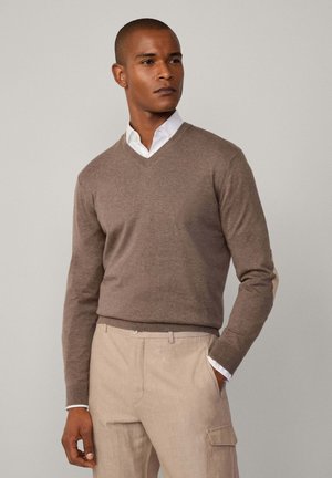 Een bruine v-hals trui gemaakt van zacht gebreid materiaal, met lange mouwen en een subtiele textuur. Gecombineerd met lichtbeige broeken en een witte blouse.