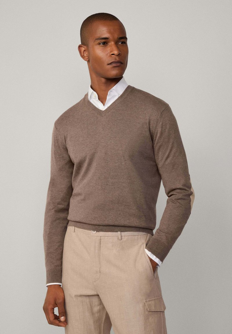 Maglione marrone a scollo a V realizzato in morbido tessuto a maglia, con maniche lunghe e una sottile texture. Abbinato a pantaloni beige chiaro e camicia bianca.