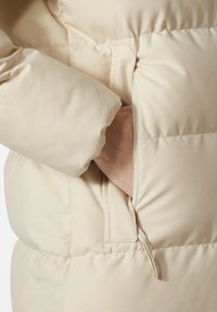 Beige puffrová bunda s měkkou, hladkou texturou; má zipovou kapsu po straně a mírně elastický manžet.