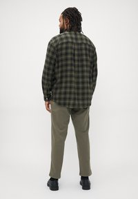 Camicia di flanella a quadretti verdi e neri, maniche lunghe, con polsini abbottonati, abbinata a pantaloni verde oliva e scarpe nere, vista da dietro.